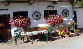 A picturesque house with large flower boxes full of colorful flowers. In front of it stands a table with chairs, surrounded by more plants.