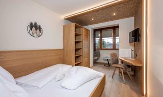 A cozy bedroom with a double bed and wooden shelves. In the background, there is a workspace with a chair and a seating area in front of a window.