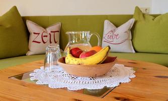 A fruit composition in a bowl with a carafe of water on a wooden table. In the background, decorative cushions can be seen.