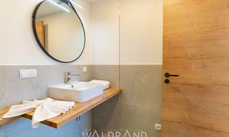 A modern bathroom with a round mirror and an elegant sink. The walls are bright, and there are wood and natural stone accents.