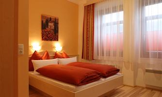 A cozy bedroom with a large bed and orange pillows. The walls are painted in a warm yellow tone and large windows let in plenty of daylight.