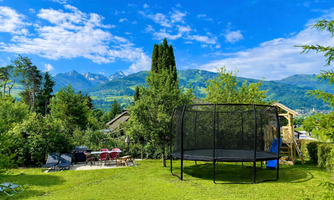 A green garden with a trampoline and a view of the mountains. In the background, trees and a small seating area can be seen.