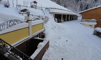 A snowy courtyard with two garages and a wooden house in the background. The landscape is surrounded by snow-covered mountains.