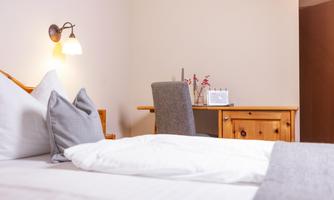 A cozy bedroom with a bed and pillows. Next to the bed is a desk with a lamp and decorative elements.