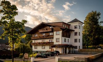 A charming multi-story building surrounded by green nature. The facade is bright, featuring balconies and a beautiful roof.