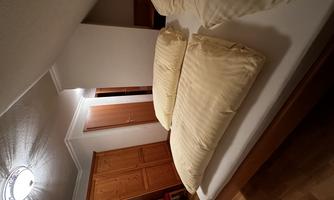 A cozy bedroom with a large bed and two pillows. The room has a wooden floor and a warmly lit ceiling.