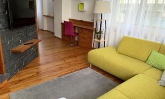 A cozy living room with a yellow sofa and wooden floor. The room is bright and modernly furnished, with a television and a desk in the background.