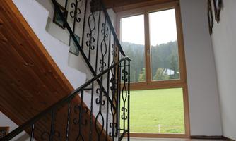A staircase with wrought iron railing and a large window front. Through the window, a green meadow and mountainous landscape are visible.