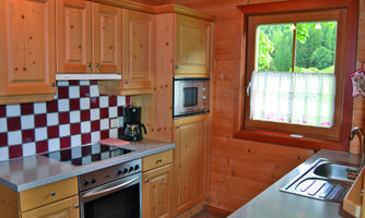 A bright kitchen with wooden ceilings and furniture. The kitchenette features modern appliances and a large window with a view of the greenery.