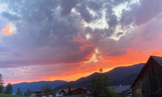 A beautiful sunset over the mountains with colorful clouds. In the foreground, barns and trees can be seen.