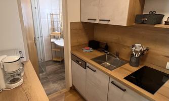 A modern kitchen made of wood and light materials. It is well-equipped and has an open connection to an adjacent room.