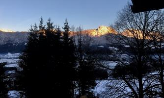 A wintry landscape with snow-covered mountains and fir trees. The sun shines gently on the peaks, creating a warm lighting mood.