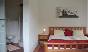 A cozy bedroom with a double bed and a nightstand. In the background, there is a bathroom with a shower and a toilet.