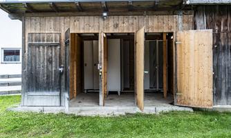 A simple wooden structure with several open doors leading to shower stalls. The floor is made of concrete, and the surroundings are landscaped with green grass.
