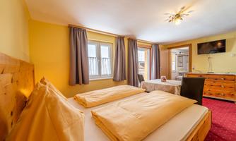 A cozy bedroom with two beds and yellow walls. Windows with curtains and a table with chairs are also visible.