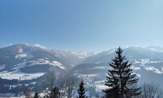 A picturesque mountain landscape with snow-covered valleys and a clear blue sky. Coniferous trees frame the view.