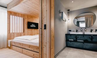 A modern room with wooden walls and a cozy bed. On the left side, there is a stylish bathroom with an elegant sink arrangement.