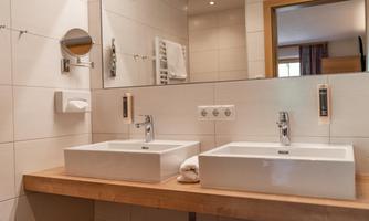 A modern bathroom with two sinks and a large mirror. The walls are tiled in light colors and the furnishings are simple and elegant.