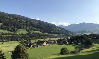A picturesque landscape with gentle hills and forested mountains. In the foreground lies a quiet village surrounded by green meadows.