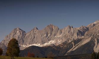 An impressive mountain landscape with snow-capped peaks and a clear sky. In the foreground, there are green meadows and some trees visible.