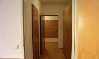 A bright hallway with wooden doors and walls. On the left side is an open room and in the background, coat hooks are visible.
