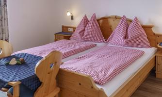 A cozy bedroom with two single beds and red-and-white checked bedspreads. The room is decorated in warm wooden tones and has a small table with chairs.
