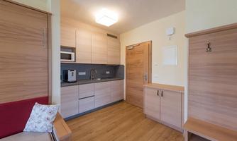 A modern small kitchen with wooden cabinets and a dining table. The decor is simple and functional, with a cozy sofa in the foreground.