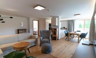 A modern living room with a bright sofa and a round table. The open space leads to a bright kitchen with large windows.