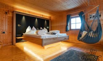 A cozy bedroom with wooden paneling and a large bed. In the corner, a hammock swings and large windows provide plenty of light.