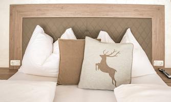 A cozy bed with white pillows and a head pillow featuring a deer motif. The headboard is made of light wood and has an elegant, padded back.