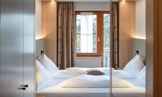 A modern bedroom with a cozy bed and wooden paneling. Large windows let in plenty of light and offer a view of nature.