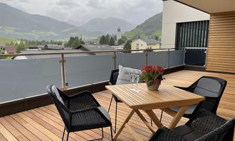 A cozy balcony with a wooden table and black chairs. In the background, green hills and a small village are visible.
