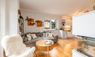A bright living room with a gray sofa, a round wooden table, and a cozy armchair. The kitchen is open and modern, with plants and decoration on the wall.