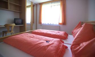 A simple room with two single beds and orange bedspreads. Next to the beds is a table and a television, the light comes through a window with curtains.