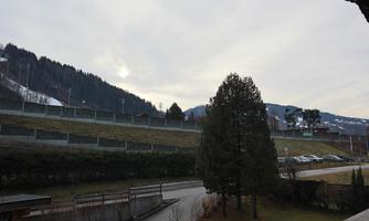 A tranquil landscape with green hills and snow-capped mountains in the background. The sky is cloudy and there are some trees near the road.