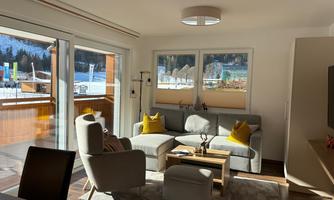 A modern living room with a comfortable sofa and soft cushions. Large windows offer a beautiful view of the mountains.