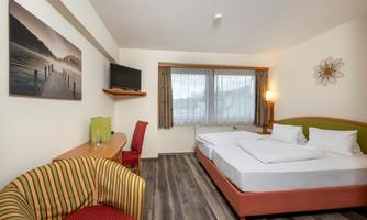 A cozy hotel room with a double bed, a television, and a small sofa. Large windows provide plenty of daylight and an inviting atmosphere.