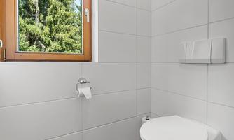A modern bathroom with white wall tiles and a simple toilet. A window offers a view of green trees outside.