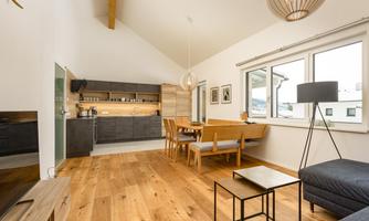 A modern living room with an open kitchen area. Large windows provide plenty of daylight, and the warm wooden ceiling creates an inviting atmosphere.