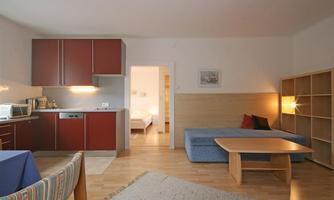 A modern living room with a kitchenette and a blue sofa. The space is bright and well-structured, with a table and chairs.