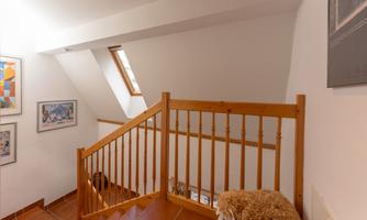 A staircase with a wooden railing leads to a bright hallway. Several pictures hang on the walls and a window lets in daylight.