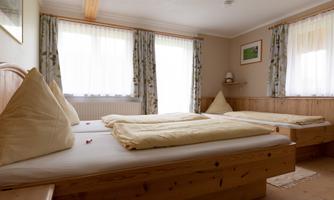 A cozy room with two beds, wooden furniture, and large windows. The windows are adorned with light curtains, and the room feels inviting and bright.