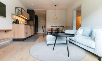 A modern living room with a cozy sofa and a round table. The room has bright colors and an open kitchen in the background.