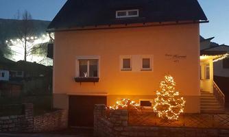 A cozy house with a warm light. In front of the house stands a decorated Christmas tree.