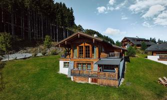 A charming wooden house on a green meadow, surrounded by trees. The sky is clear and there are a few clouds.