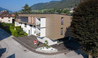 A modern residential house with clean lines and large windows. It is situated in a green environment with mountains in the background.