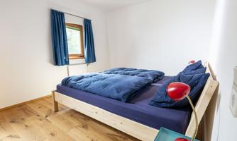 A modern bedroom with a light wooden floor. The bed is decorated with blue bedspreads and pillows, and there is a nightstand with a red lamp.