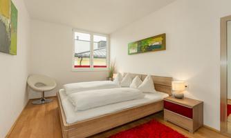 A modern bedroom with a double bed and white bedding. There is a red carpet and a cozy sitting area with a chair.