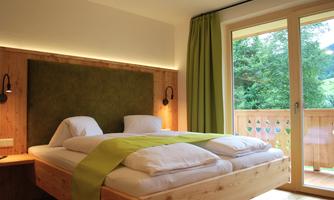 A modern bedroom with a large bed and green cushions. The windows are open and offer a view of nature outside.