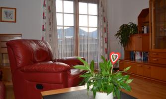 A cozy living room with a red armchair and plants on a table. Lots of light comes in through the windows and there is a view of the mountains outside.